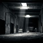 A haunting black and white photo of an abandoned warehouse with a solitary chair illuminated by sunlight through a window.