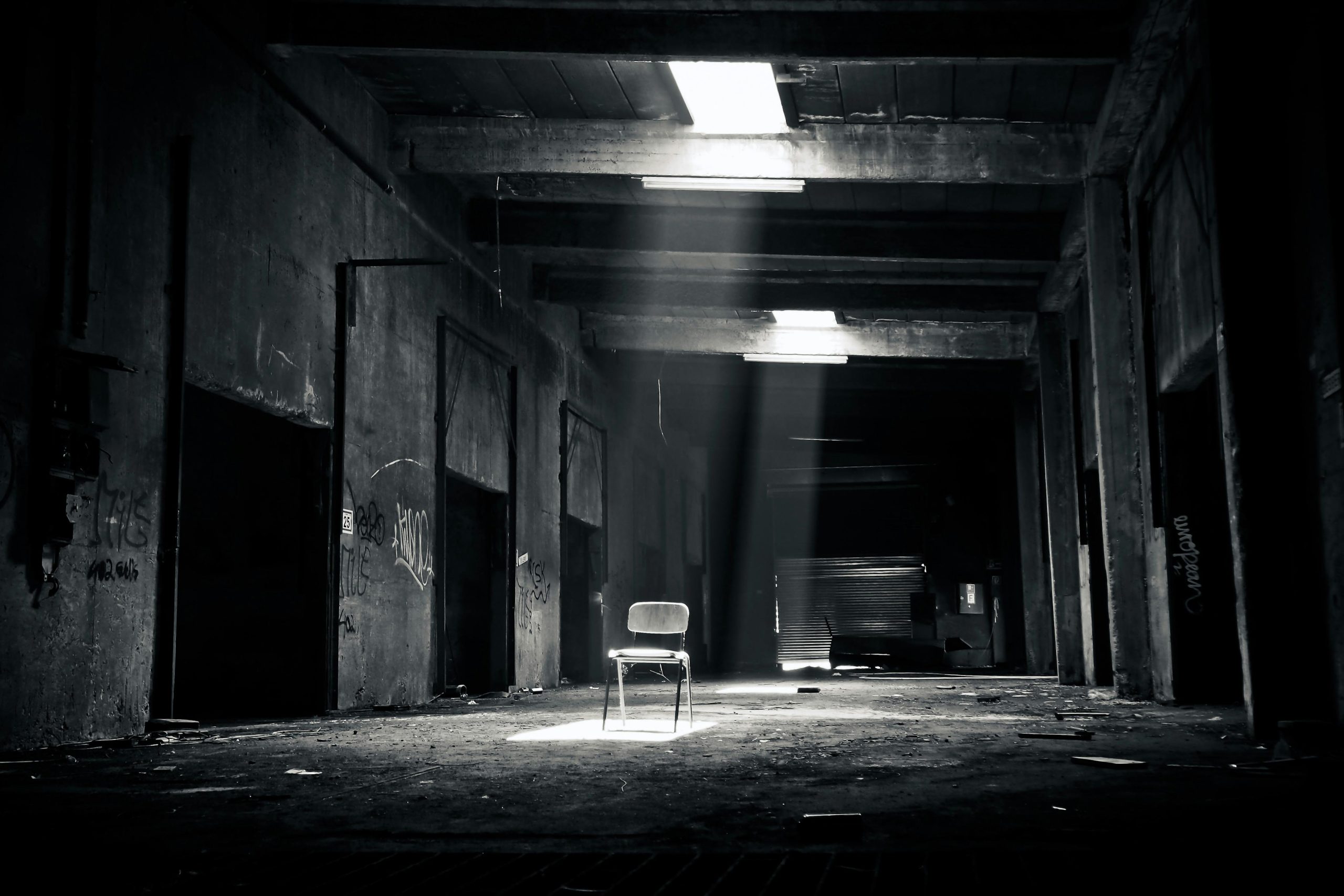 A haunting black and white photo of an abandoned warehouse with a solitary chair illuminated by sunlight through a window.